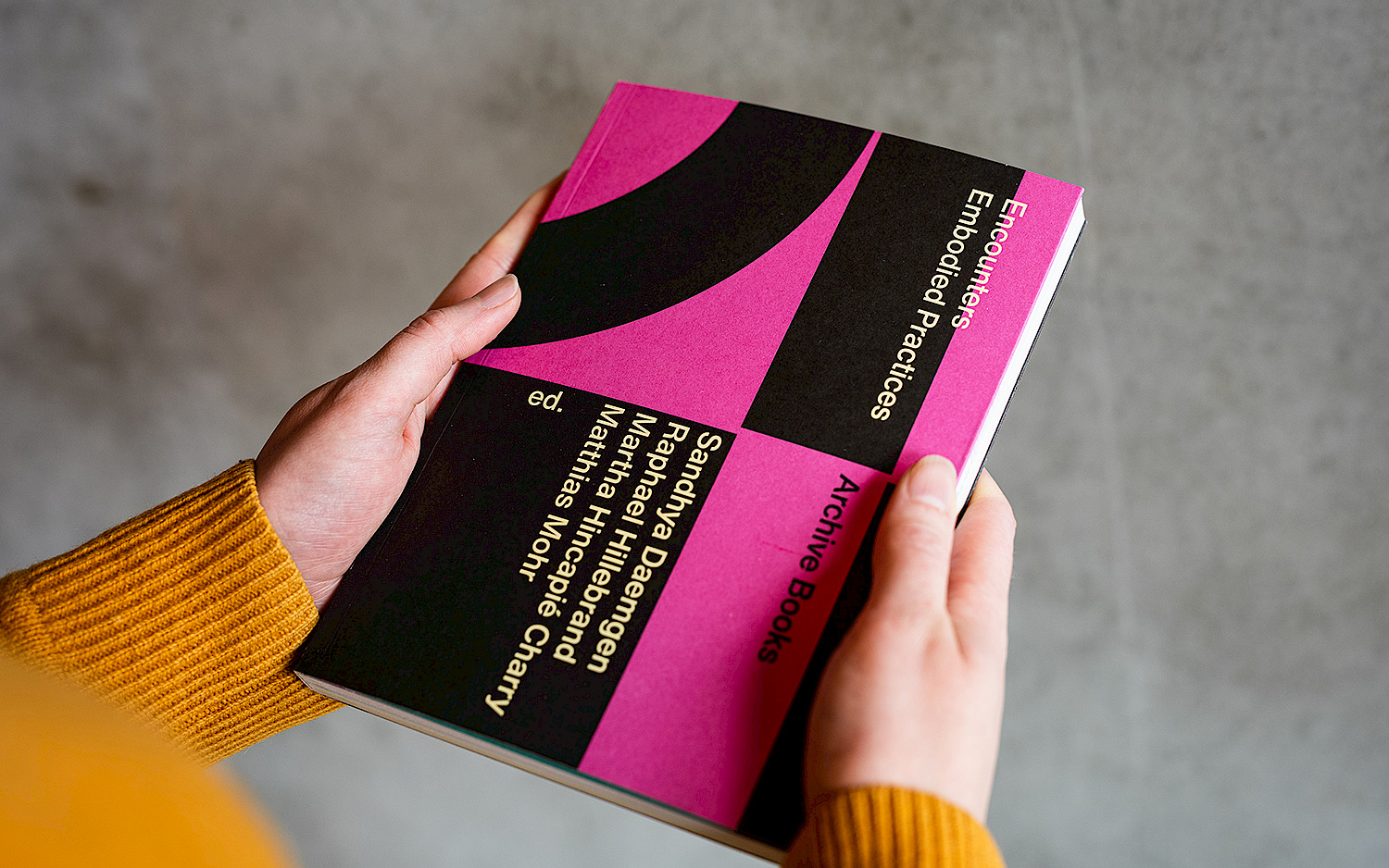Eine Person hält das Buch "Encounters – Embodied Practices" in der Hand, das Cover ist grafisch gestaltet und schwarz und pink.