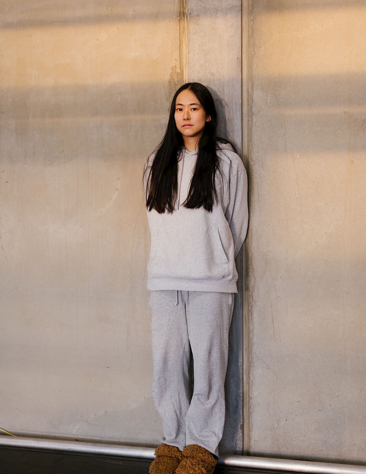 Phil Dera Hanako Hayakawa in grauer Jogginghose und grauem Pullover vor einer grauen Betonwand.