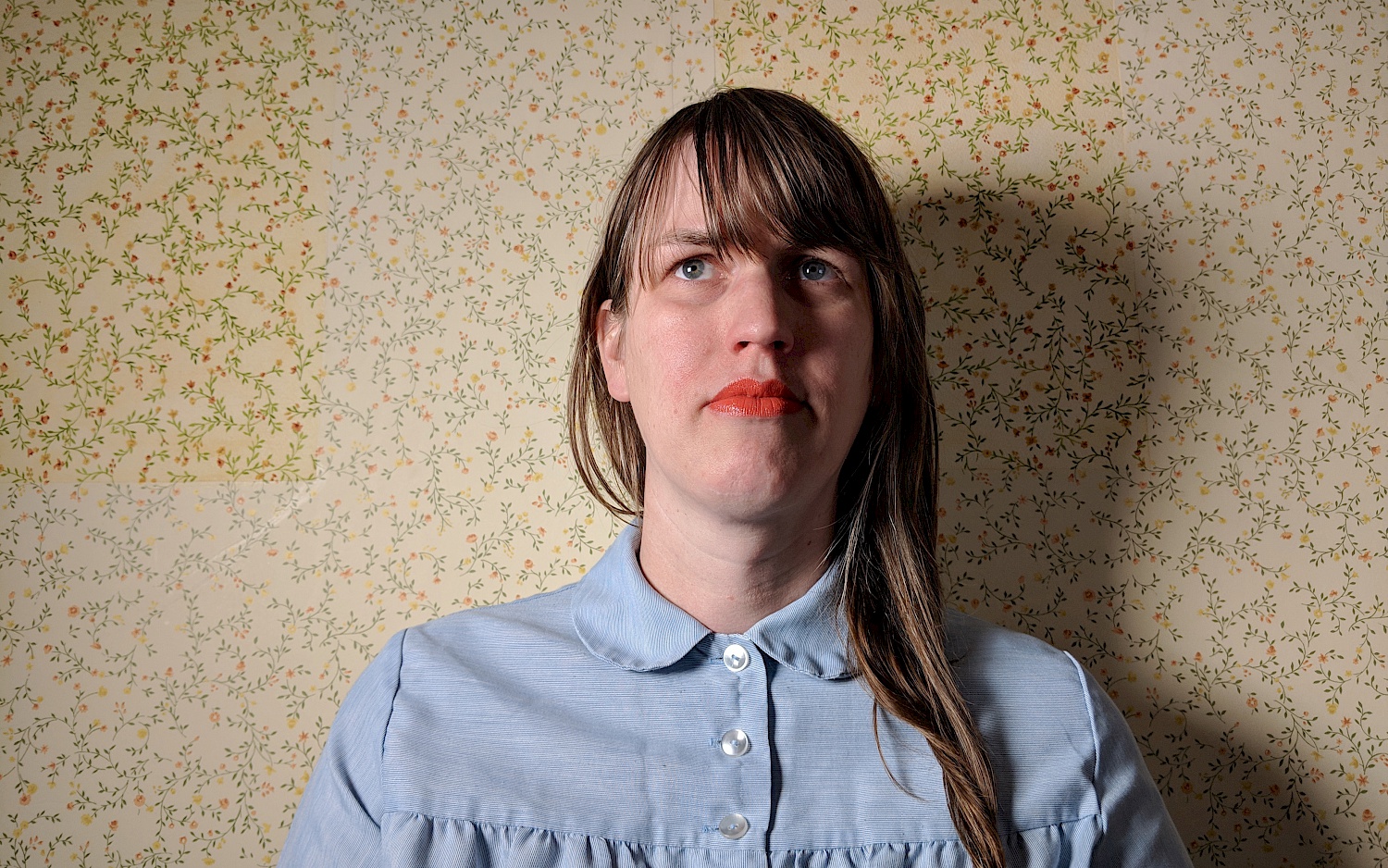 Portrait von Sarah Hennies vor einer gelb gemusterten Tapete. Sie trägt roten Lippenstift und eine hellblaue Bluse.