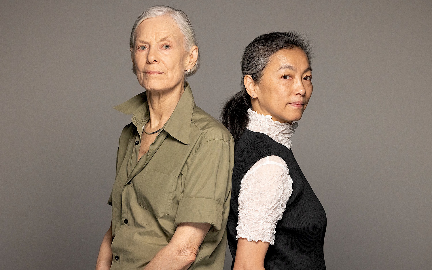 Two people stand back to back and look sideways into the camera.