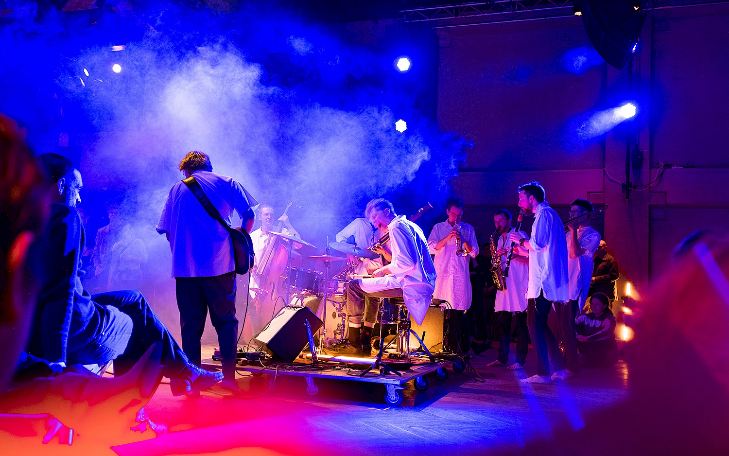 Musiker*innen spielen in blau-rotem Licht und Nebel. Intensive Atmosphäre, verschiedene Instrumente, Publikum sitzt und steht nah dabei.