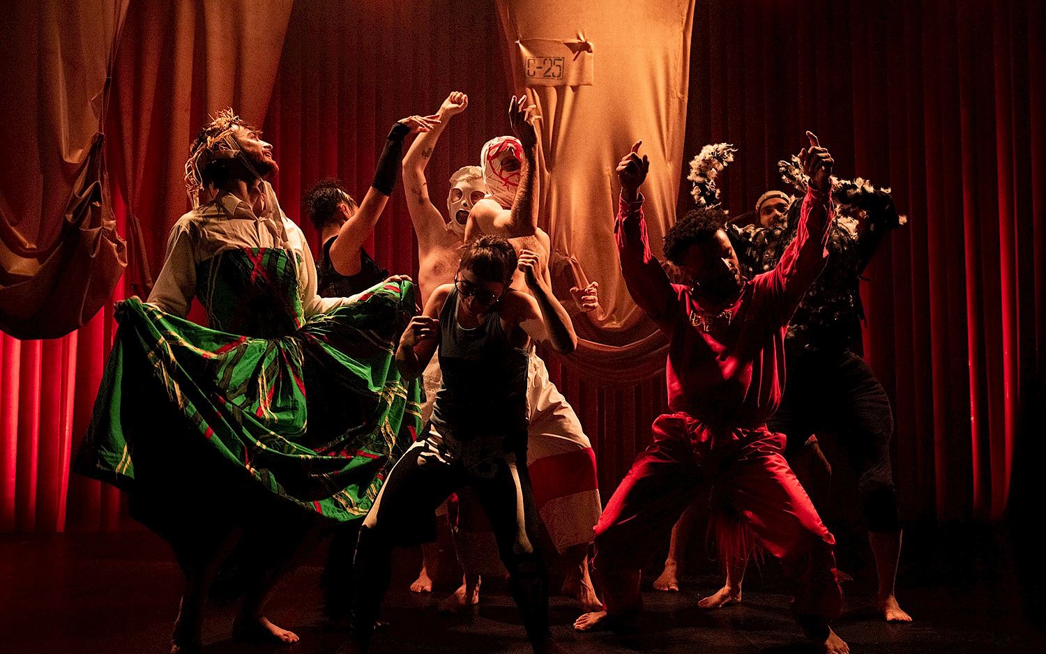 Group of performers in expressive poses on stage with red curtains, wearing imaginative costumes and dramatic lighting.