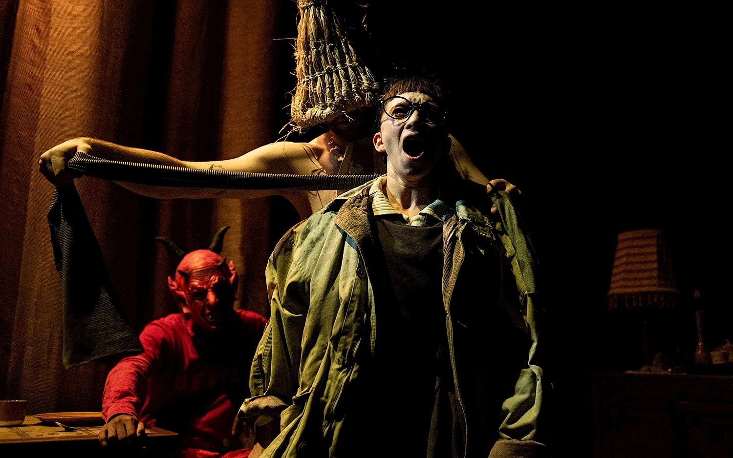 Theatrical scene with a person screaming in the foreground, a performer with a straw hat behind them, and a devil-costumed figure in the background under moody stage lighting.