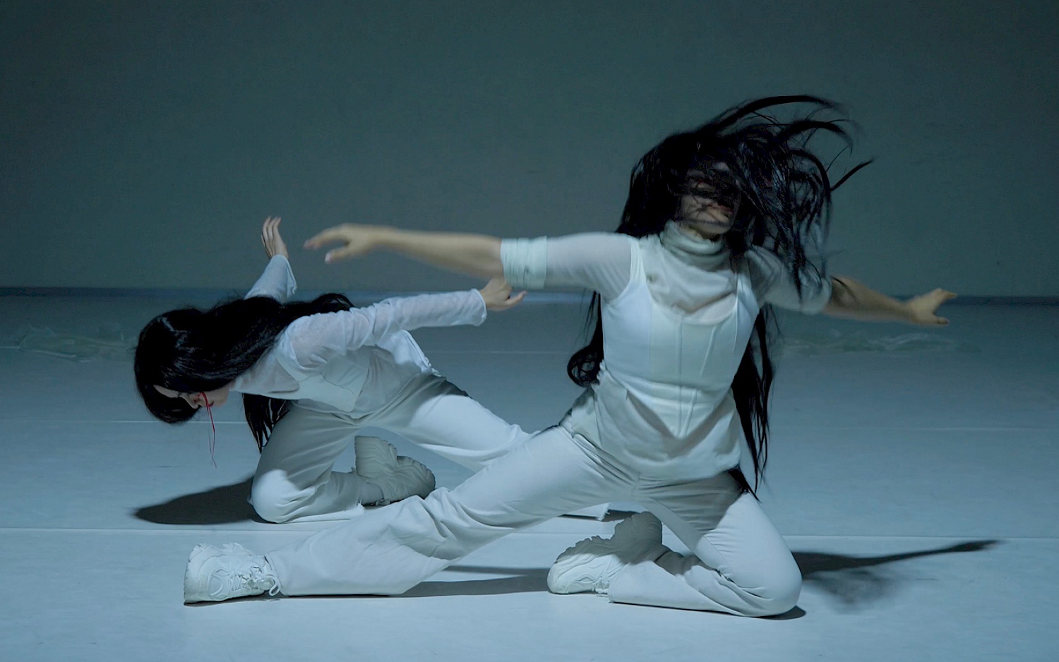 Performance excerpt from Orfeo by Sasha Amaya. Two dancers in white outfits. One is kneeling on the ground with their head bowed, while the other strikes a dramatic pose with outstretched arms and hair flying.