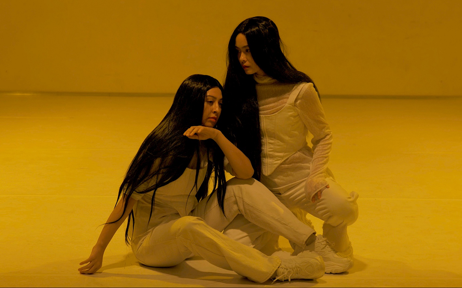 Performance-Ausschnitt aus "Orfeo". Zwei Performerinnen, weiß gekleidet mit schwarzen, langen Perücken sitzen und knien auf weiße Tanzboden in gelbem Licht.