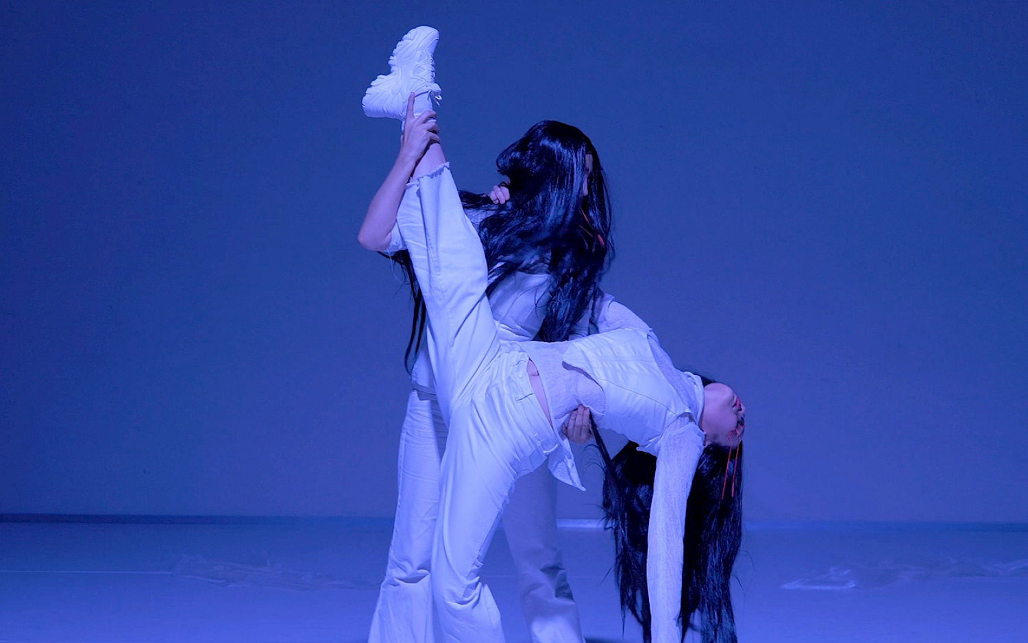 Performance-Ausschnitt von "Orfeo". In weiß gekleidete Performerinnen mit langhaarigen, schwarzen Perücken vor blauem Hintergrund. Die eine Person hält die andere mit gestrecktem Bein.