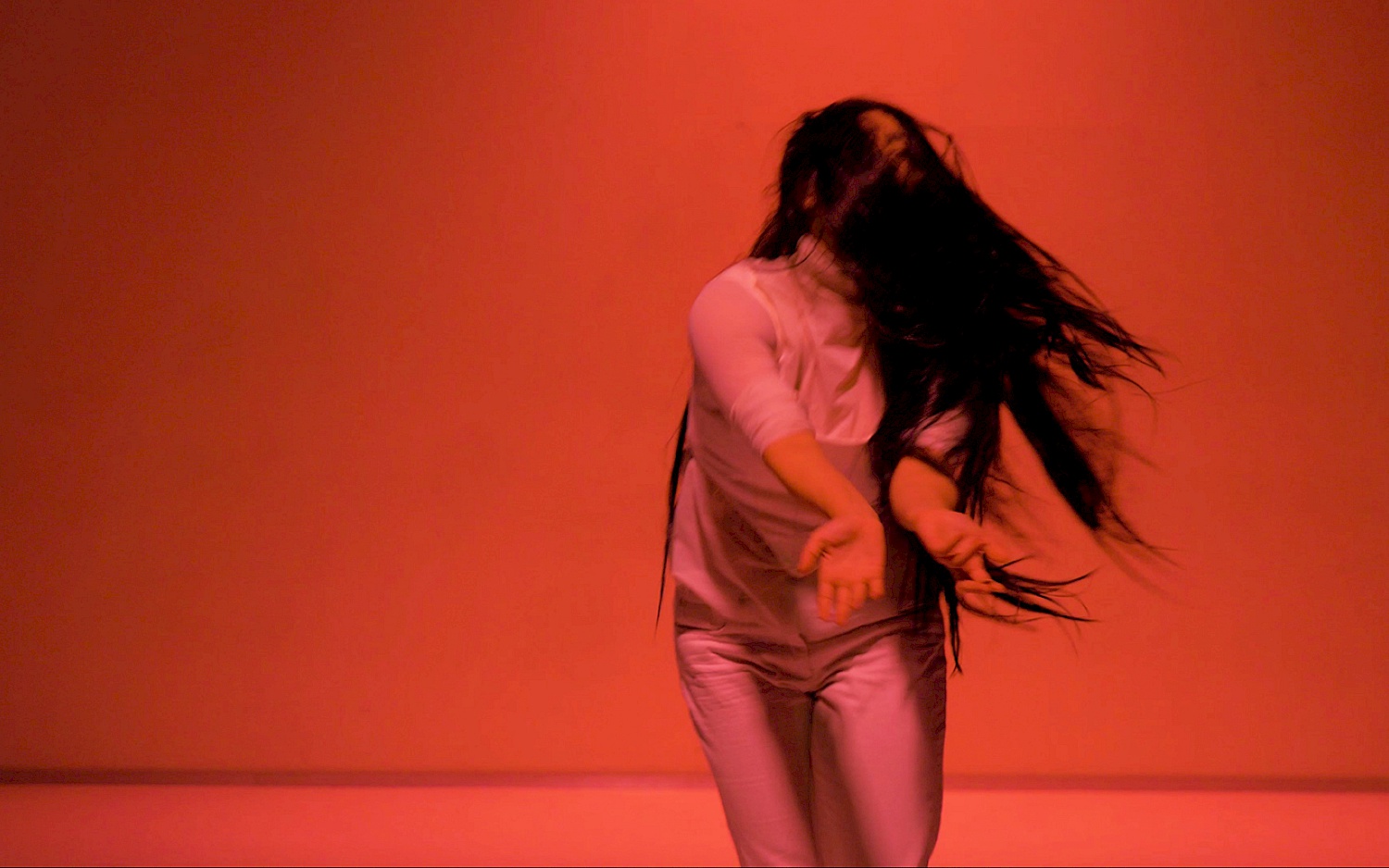 Performance-Ausschnitt aus "Orfeo". Eine Performerin in weißer Kleidung und langer, schwarzer Perücke bewegt sich in rotem Licht.