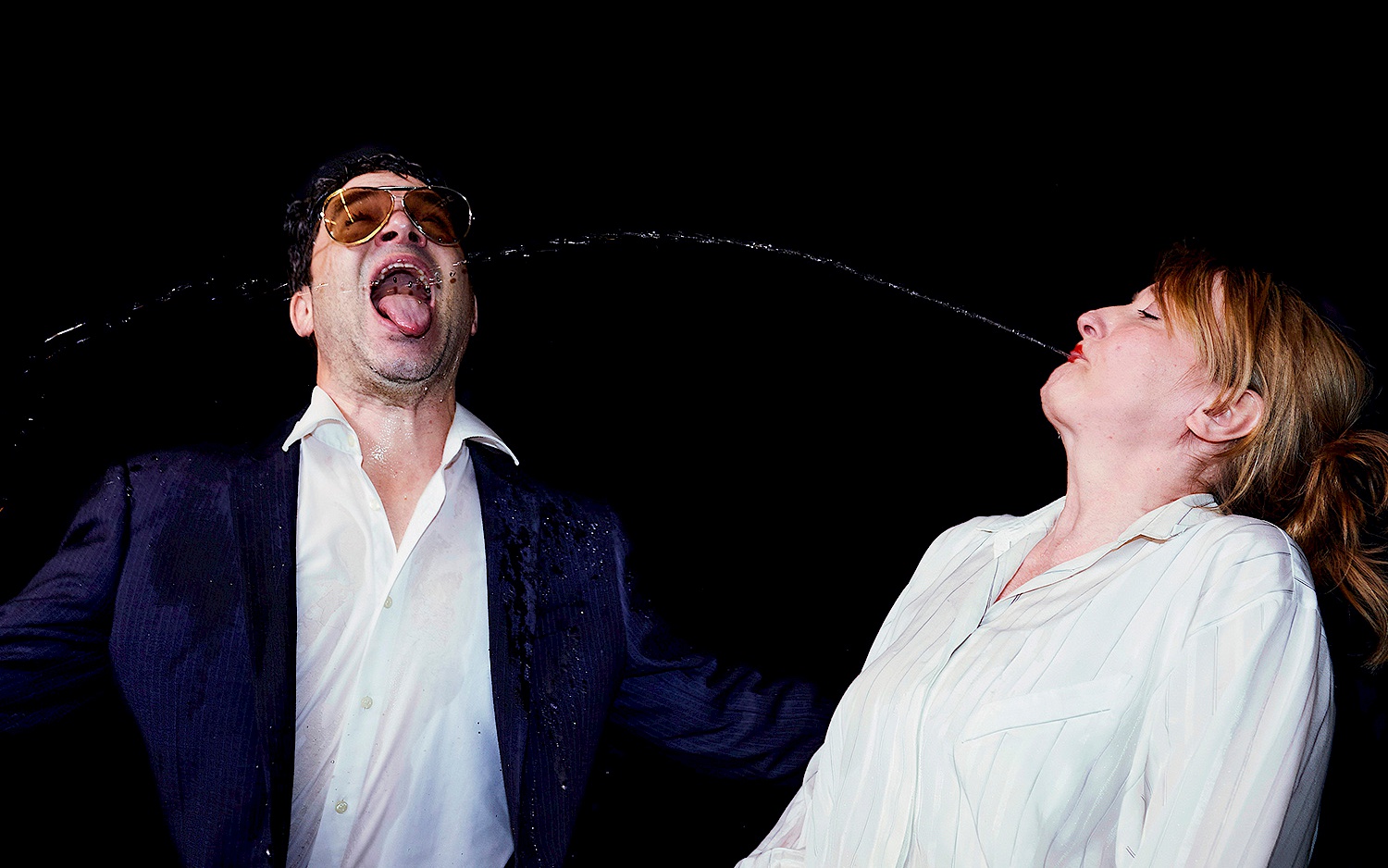 Zwei Personen in schicken Klamotten. Die rechte Person spuckt der linken im Bogen eine Flüssigkeit in den Mund, die andere Person fängt diese mit offenem Mund.