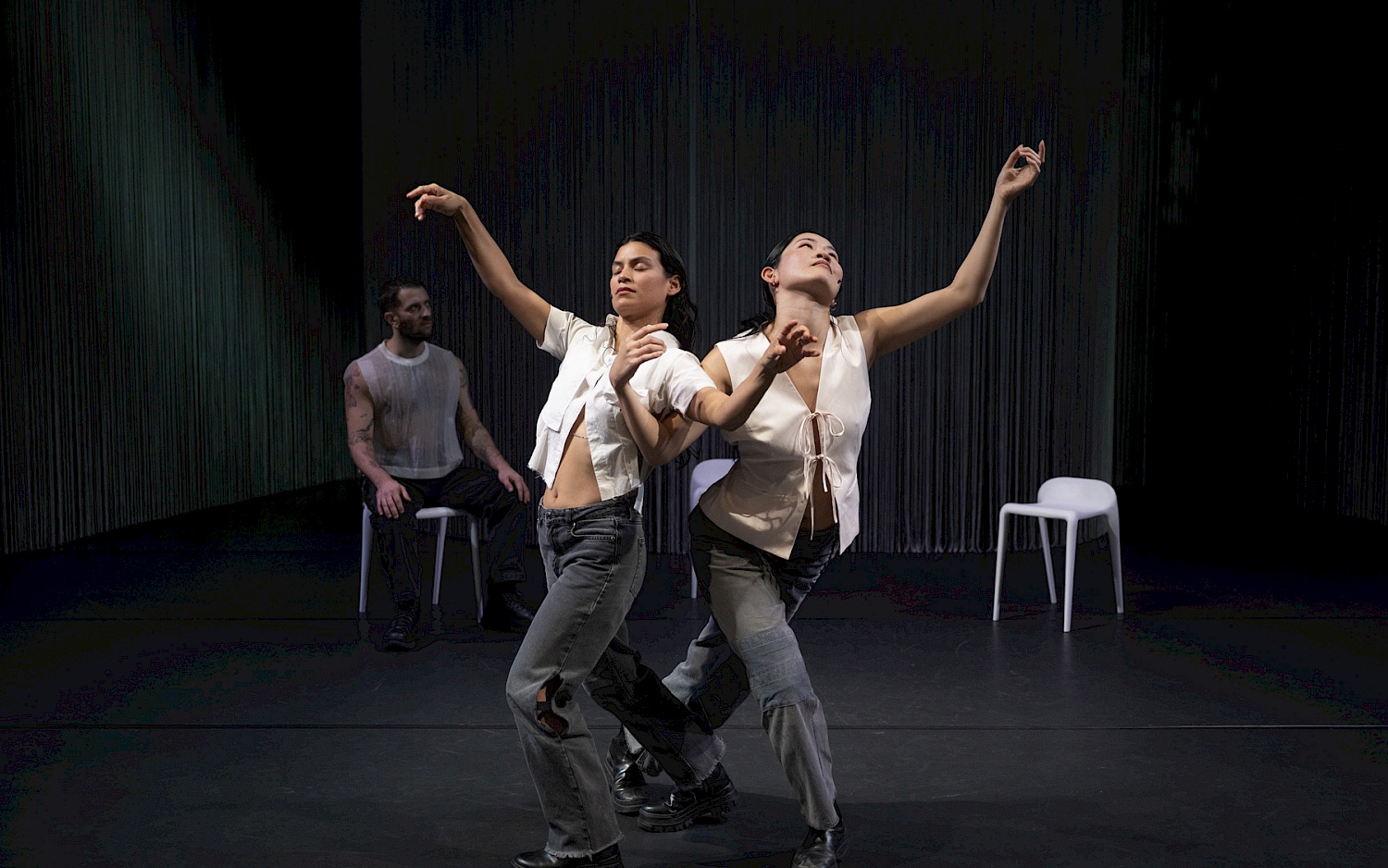 Zwei Tänzerinnen bewegen sich synchron, ein Tänzer sitzt im Hintergrund.