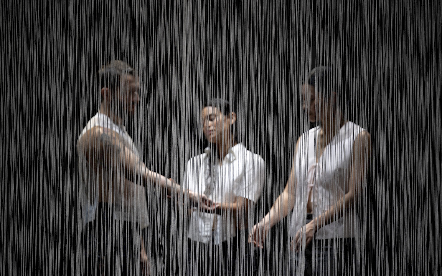 Drei Performer stehen hinter einem durchlässigen Fadenvorhang.