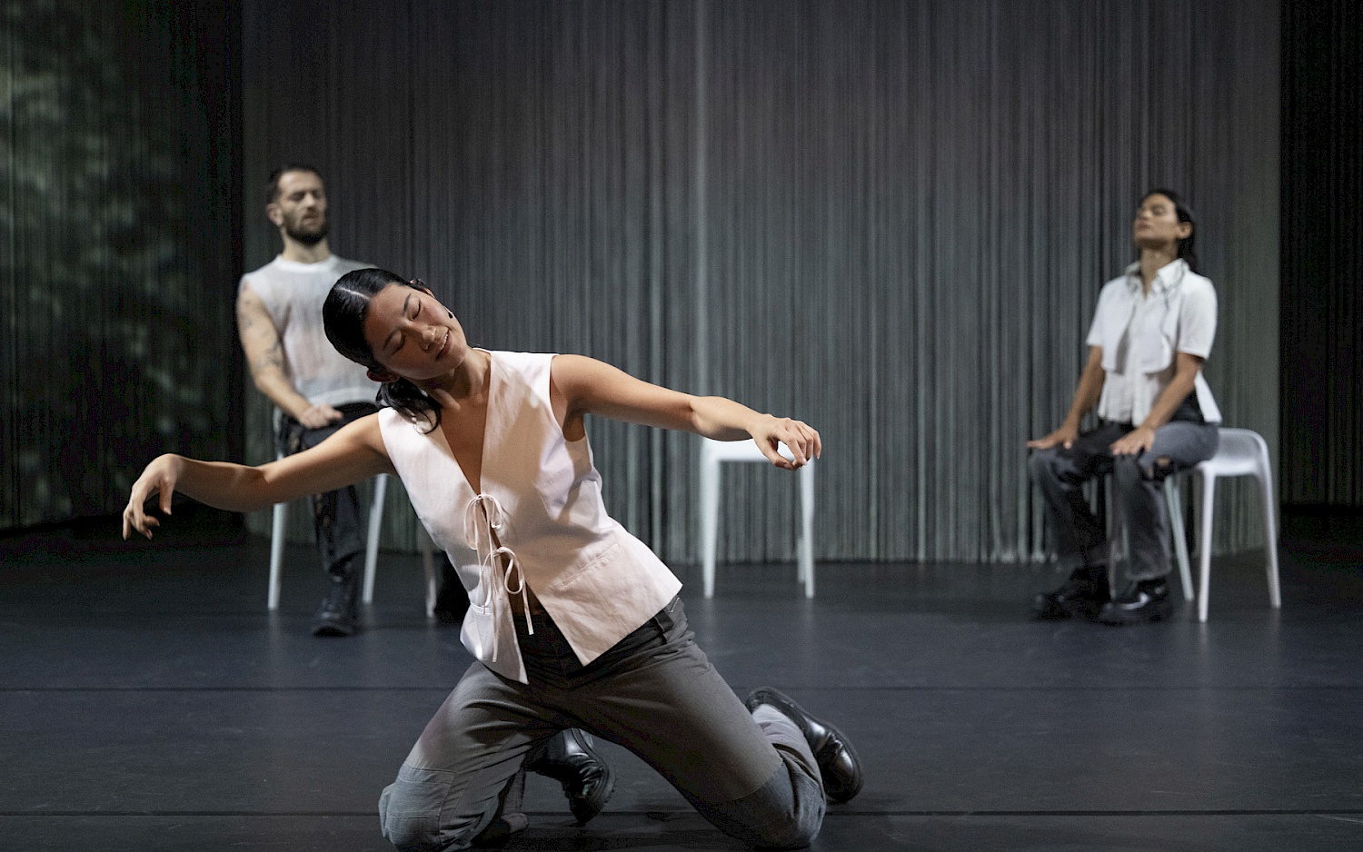 Eine Tänzerin kniet im Vordergrund mit seitlich erhobeben Armen, zwei Performer sitzen im Hintergrund.