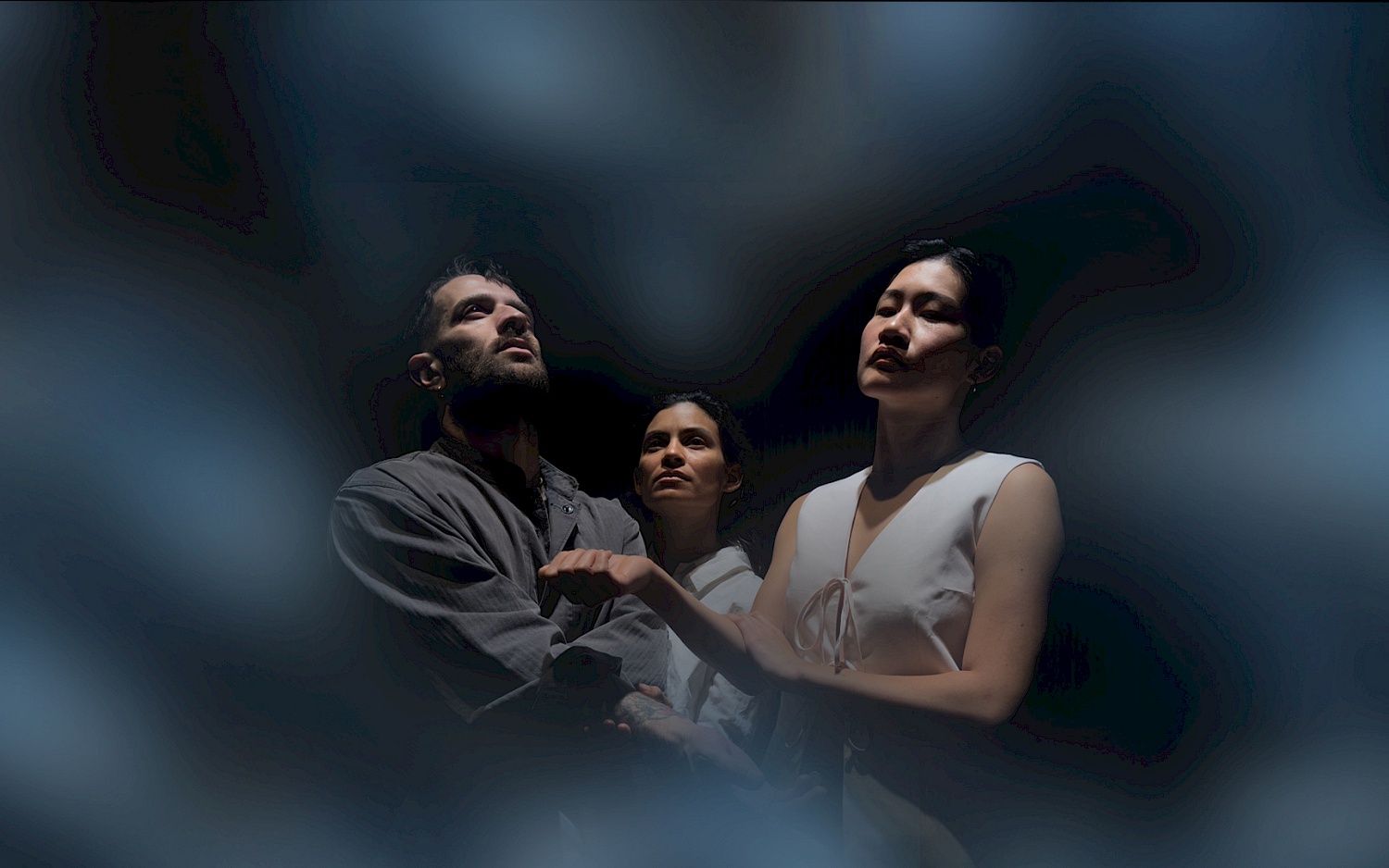 Drei Personen mit gehobenem Blick stehen eng beieinander. Um sie herum Nebel und Dunkelheit