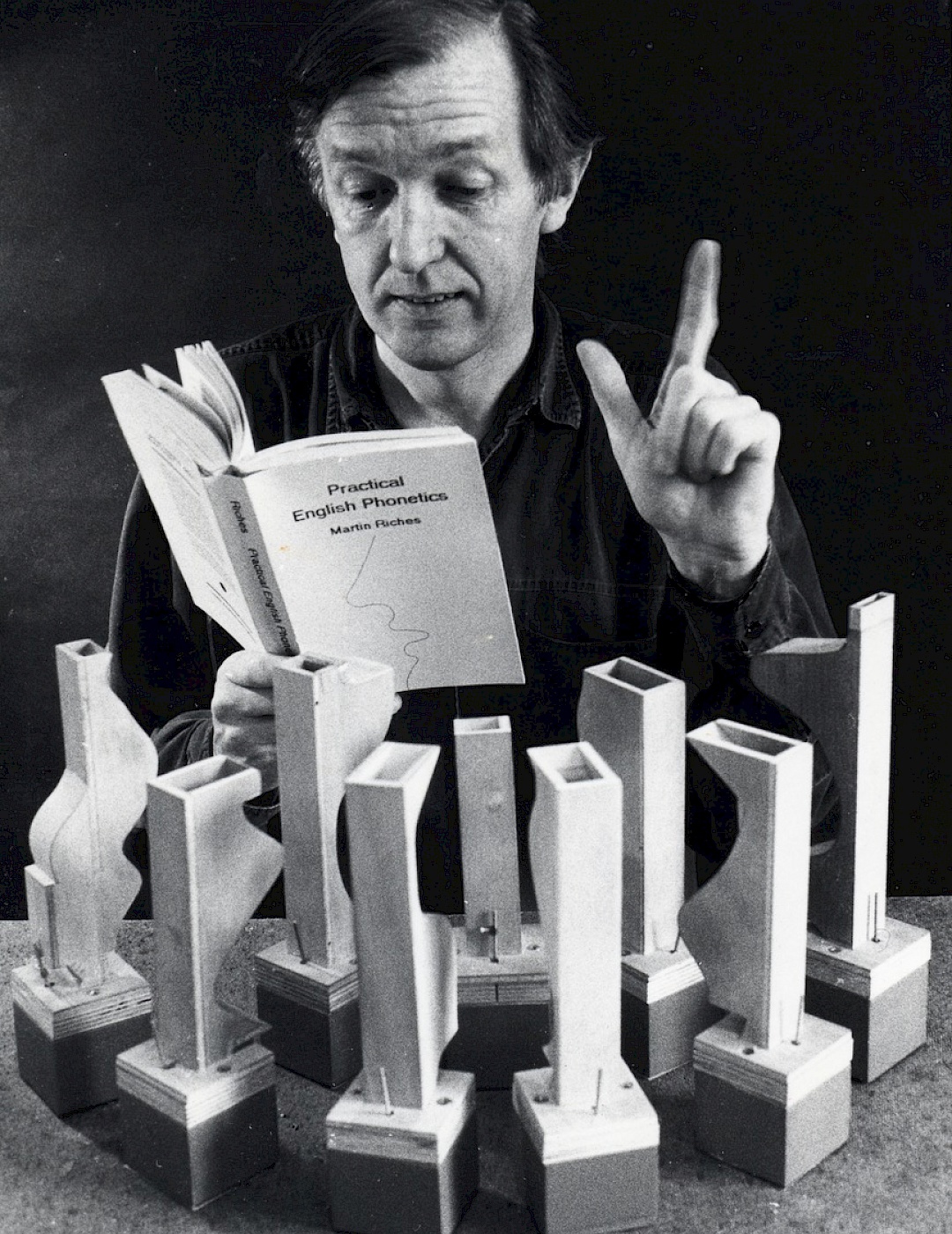 Martin Riches Black and white photograph of Martin Riches. He has his index finger raised and is reading a book. Several wooden objects are arranged in a semicircle in front of him.