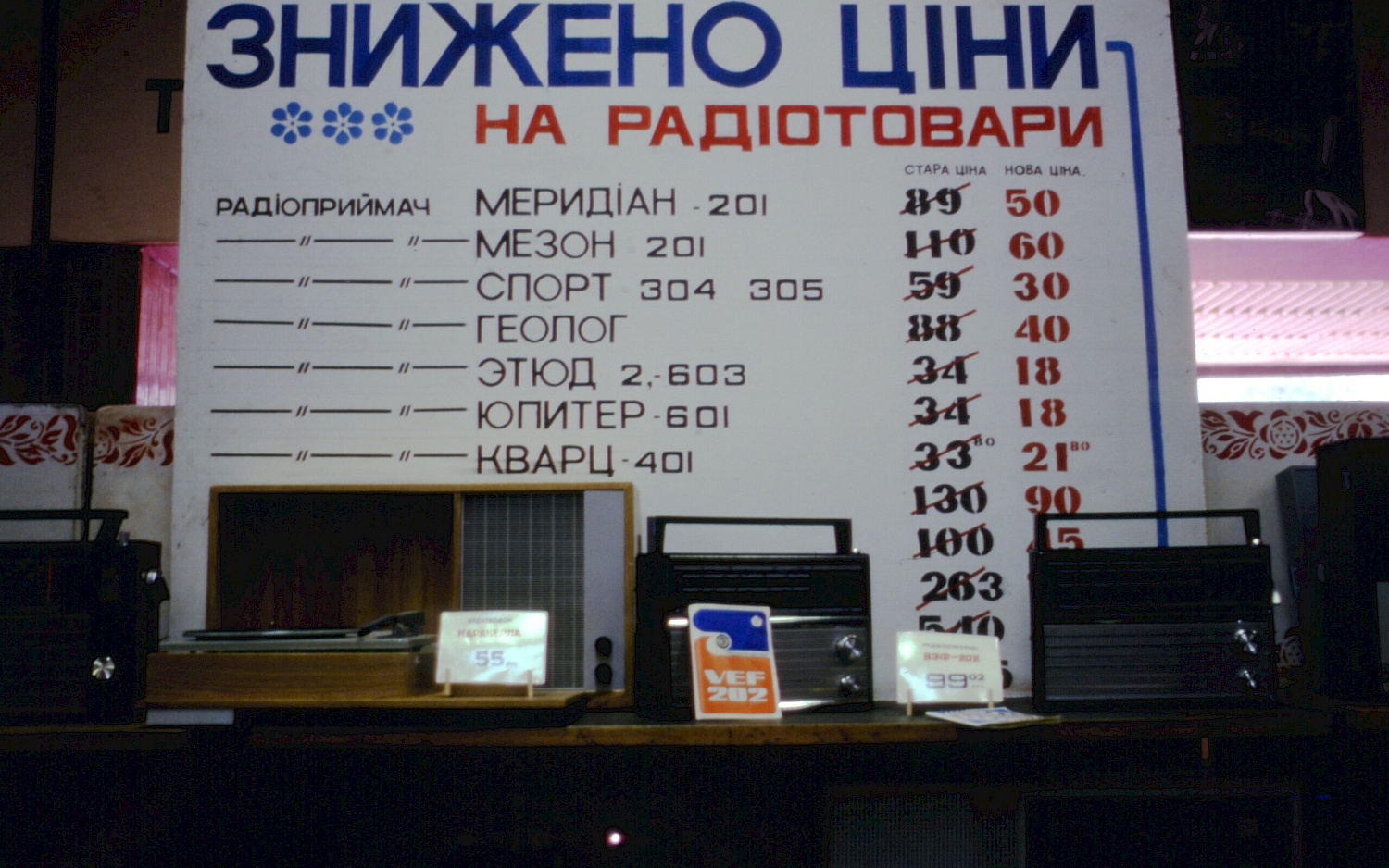 This photo features a sign in Ukrainian announcing lower prices on radios. The samples  are two VEF-202 (or its sister models, mfg. since 1971) and the Caravella tube record  player