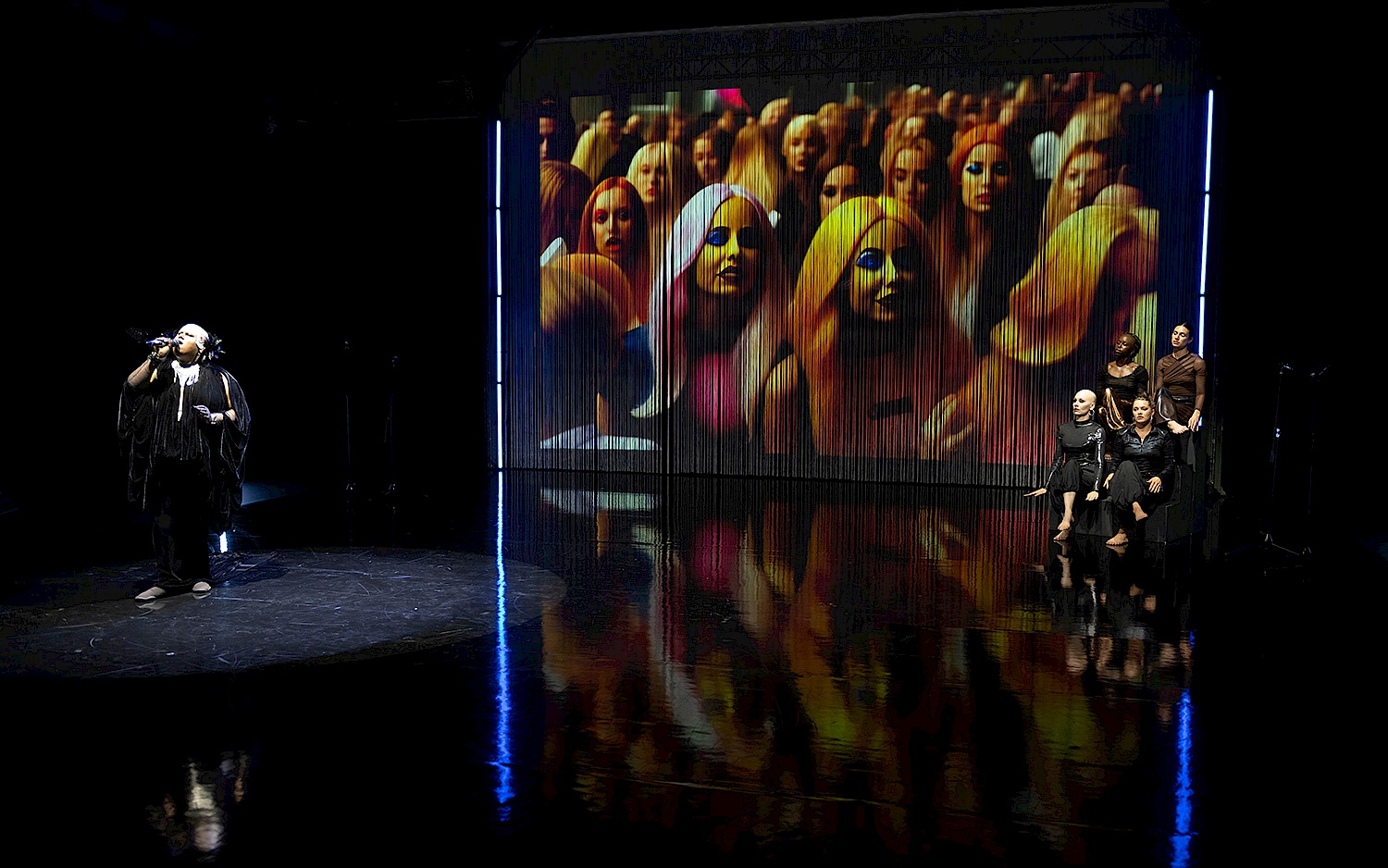 Singende Person im Spotlight vor einer Leinwand, die eine Menge an gestylten Köpfen zeigt. Rechts im Bild eine Gruppe von vier Menschen.