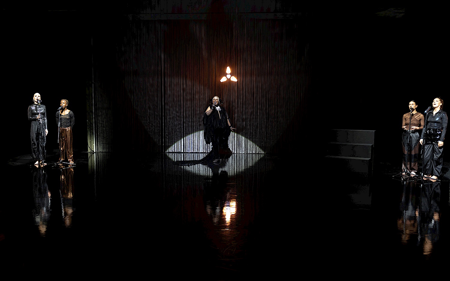Fünf singende Personen in drei Gruppen, jeweils mit Spotlight erleuchtet. Im Hintergrund lässt sich eine Leinwand aus dünnen Fäden erahnen.