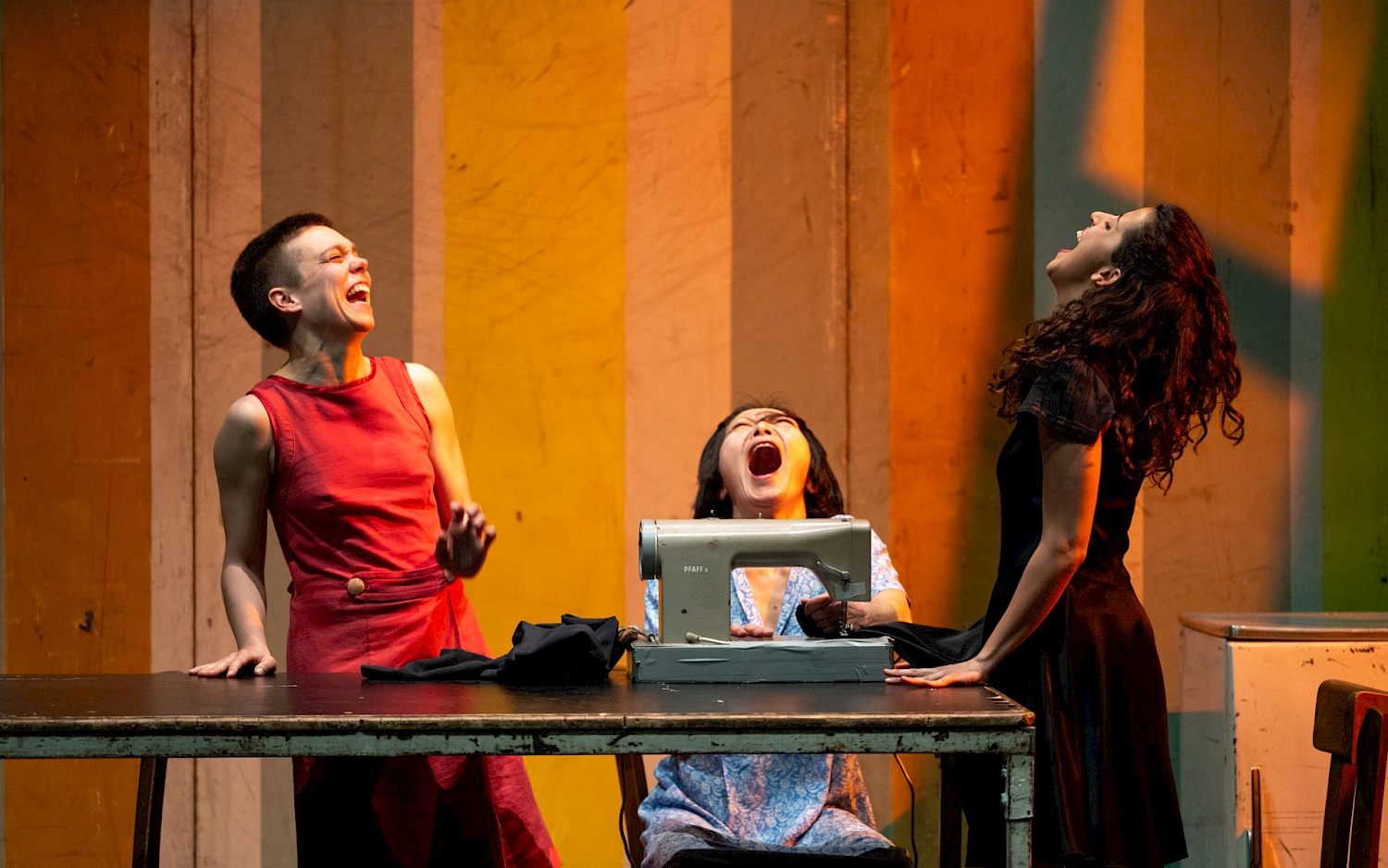 Drei Performer*innen sind um eine Nähmaschine versammelt und schreien mit erhobenen Köpfen und geschlossenen Augen.