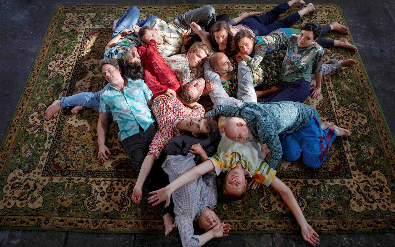 Gruppenbild der Mitglieder des ensemble mosaik: alle Personen liegen über- und untereinander auf einem großen Teppich