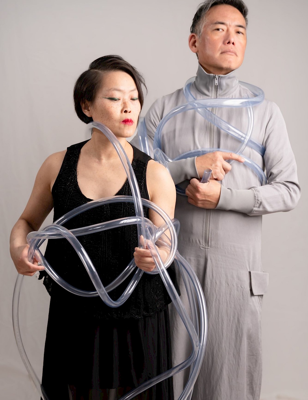 David Beecroft A woman in a black dress and a man in a gray suit are standing in front of a gray wall. A long transparent tube is partially wrapped around them.