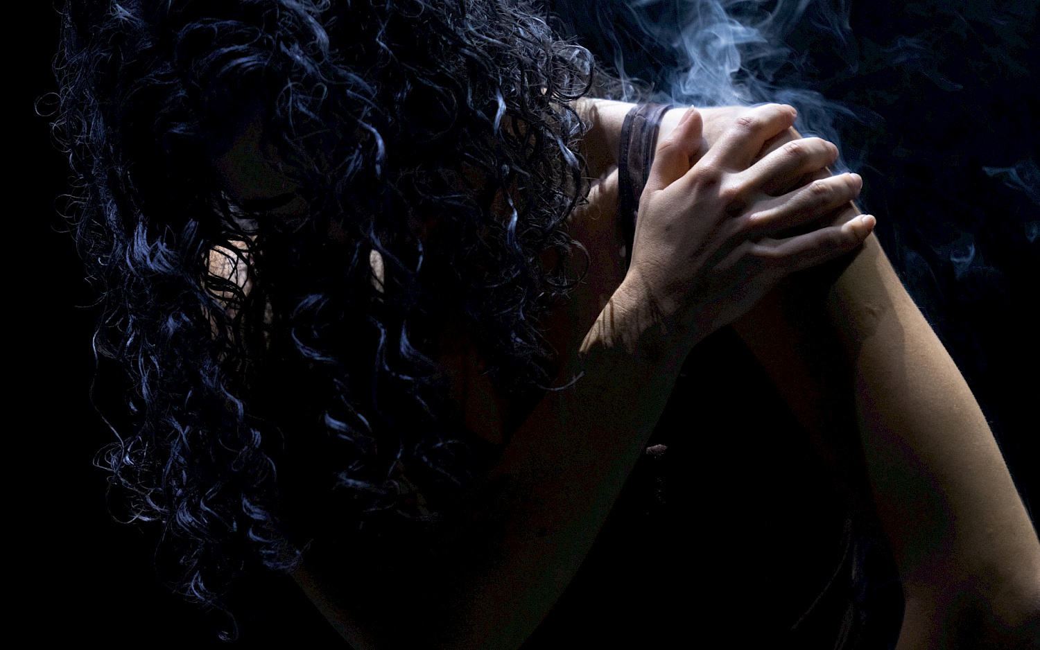 A person with long black curled  hair touches their shoulder with their hand. The affected area is smoking.