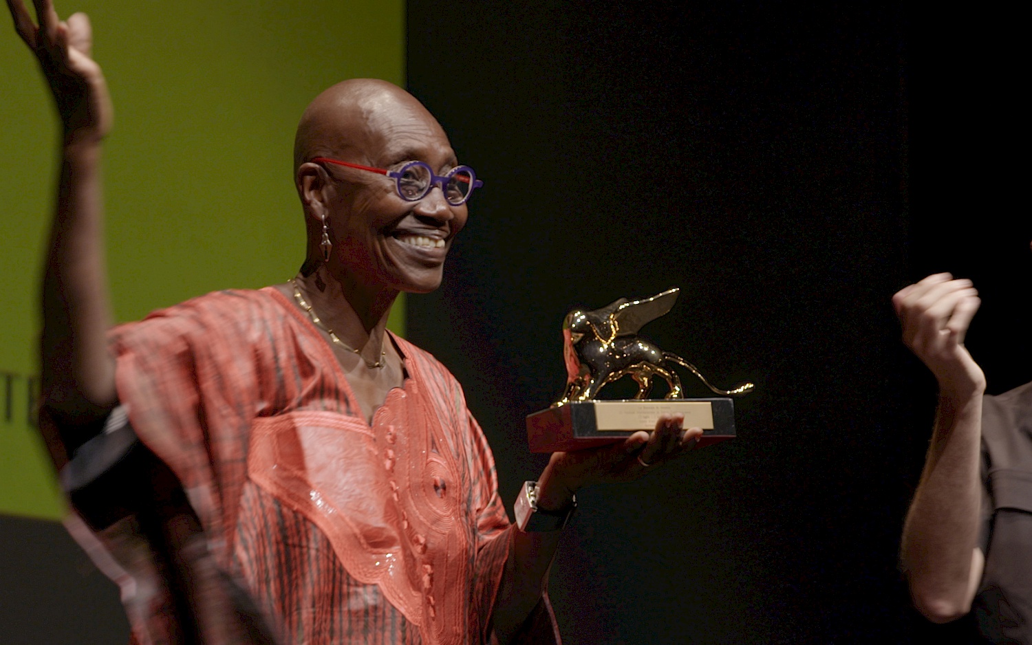 CALA FILM Film still: Germaine Acogny smiles as she holds the Golden Lion of the Venice Dance Biennale in her hand and waves to the audience.