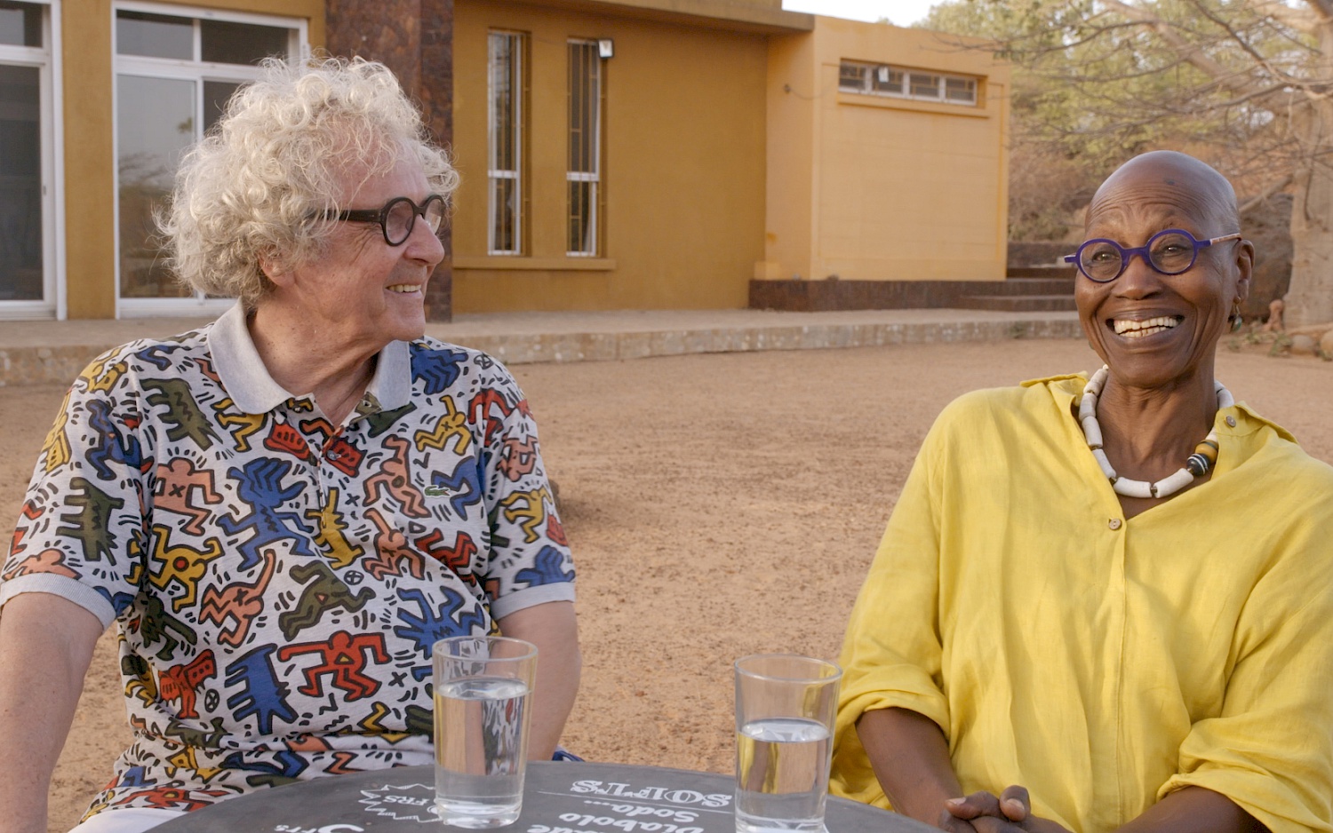 CALA FILM Film still: Germaine Acogny and Helmut Vogt sit laughing at a table in colourful clothes, in front of two glasses of water.