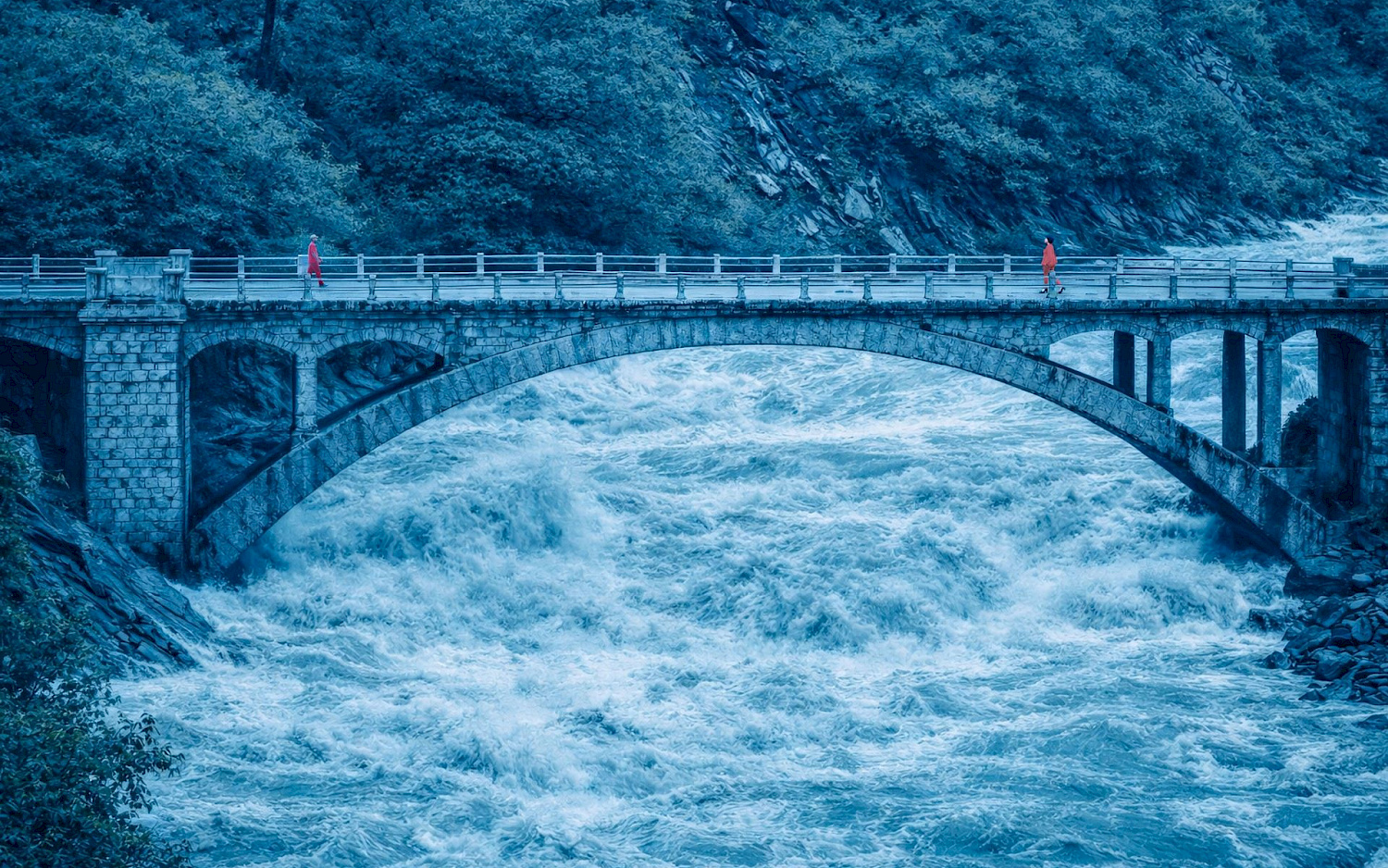 Two people are walking towards each other on a bridge that spans a raging river.