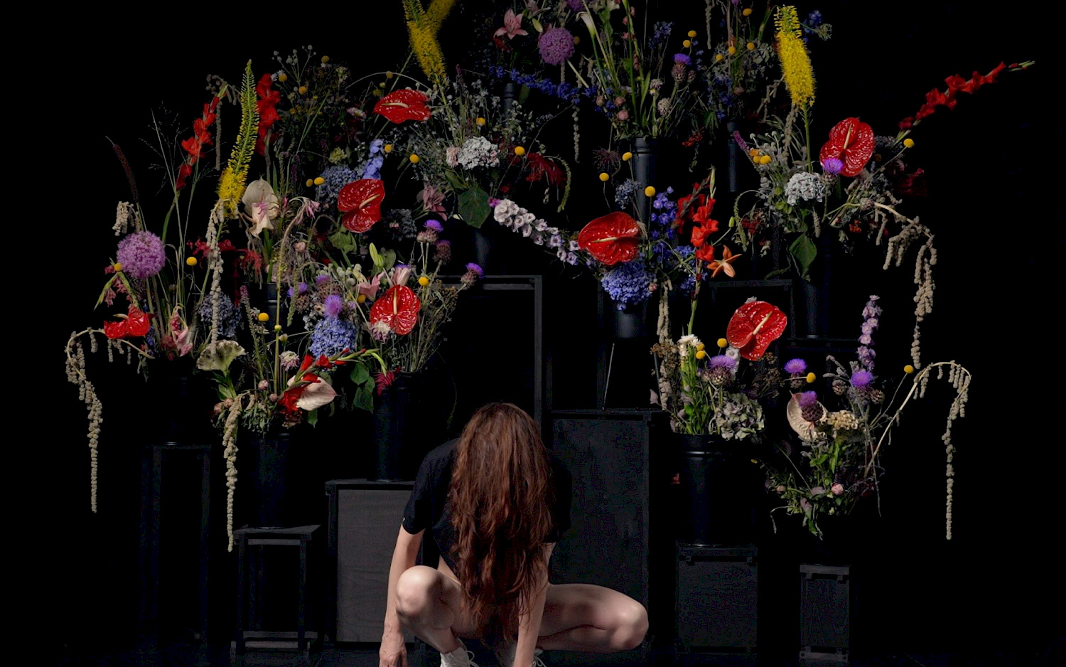 Daniel K. B. Schmidt A person is kneeling with their head bowed in front of an elaborate, colourful floral arrangement.