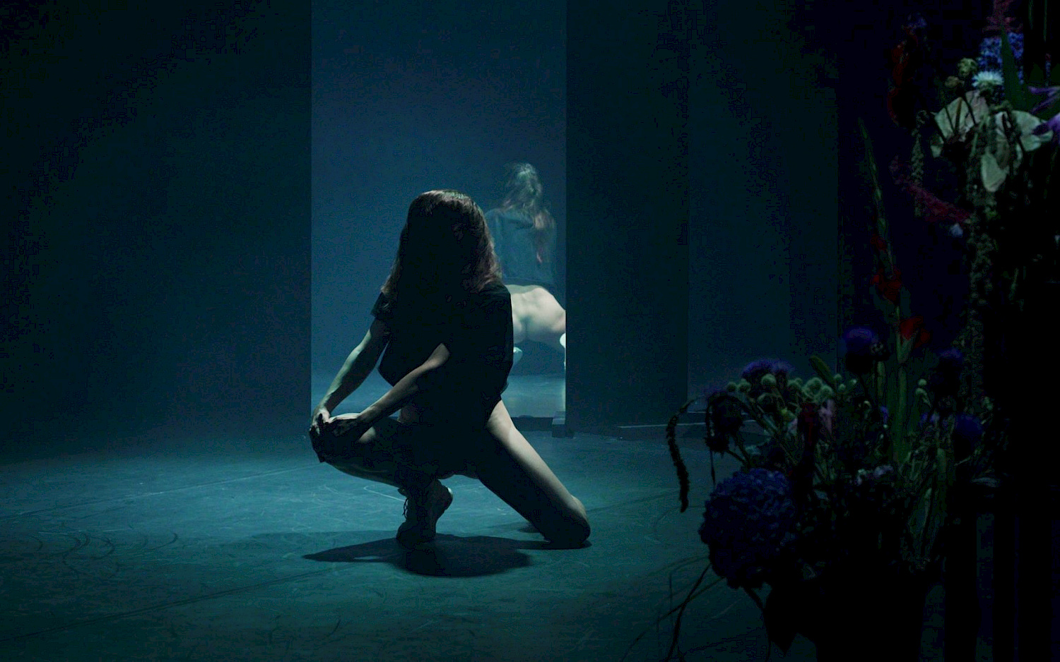 Daniel K. B. Schmidt A person is kneeling on a dark stage with their back to a large mirror. Blue flowers lie beside them.