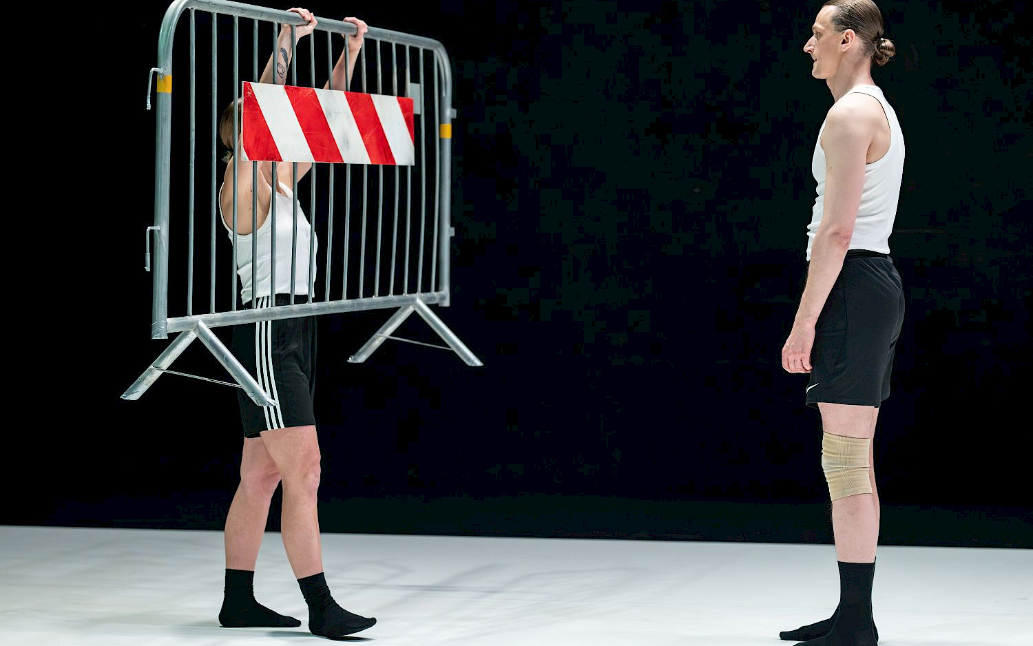 D. Alisauskas Two performers against a black background; one is holding up a barrier fence.