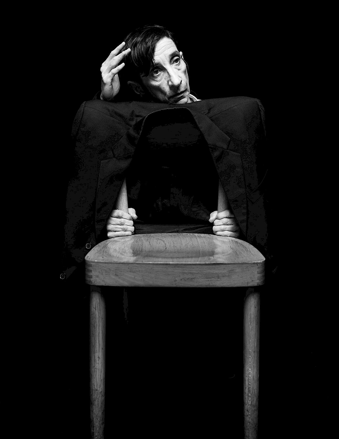 Phil Dera A black-and-white photograph of a person sitting behind a chair, looking pensive.