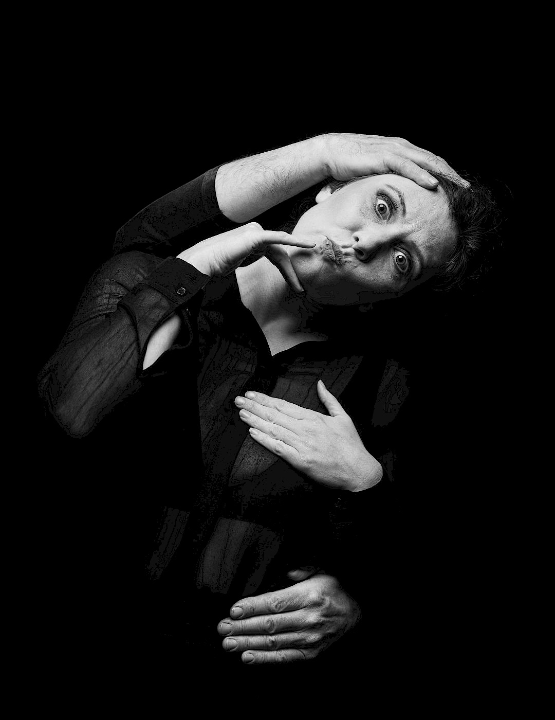 Phil Dera A black-and-white photo of a person in black clothes, with their arms wrapped around them from behind, pulling a face.
