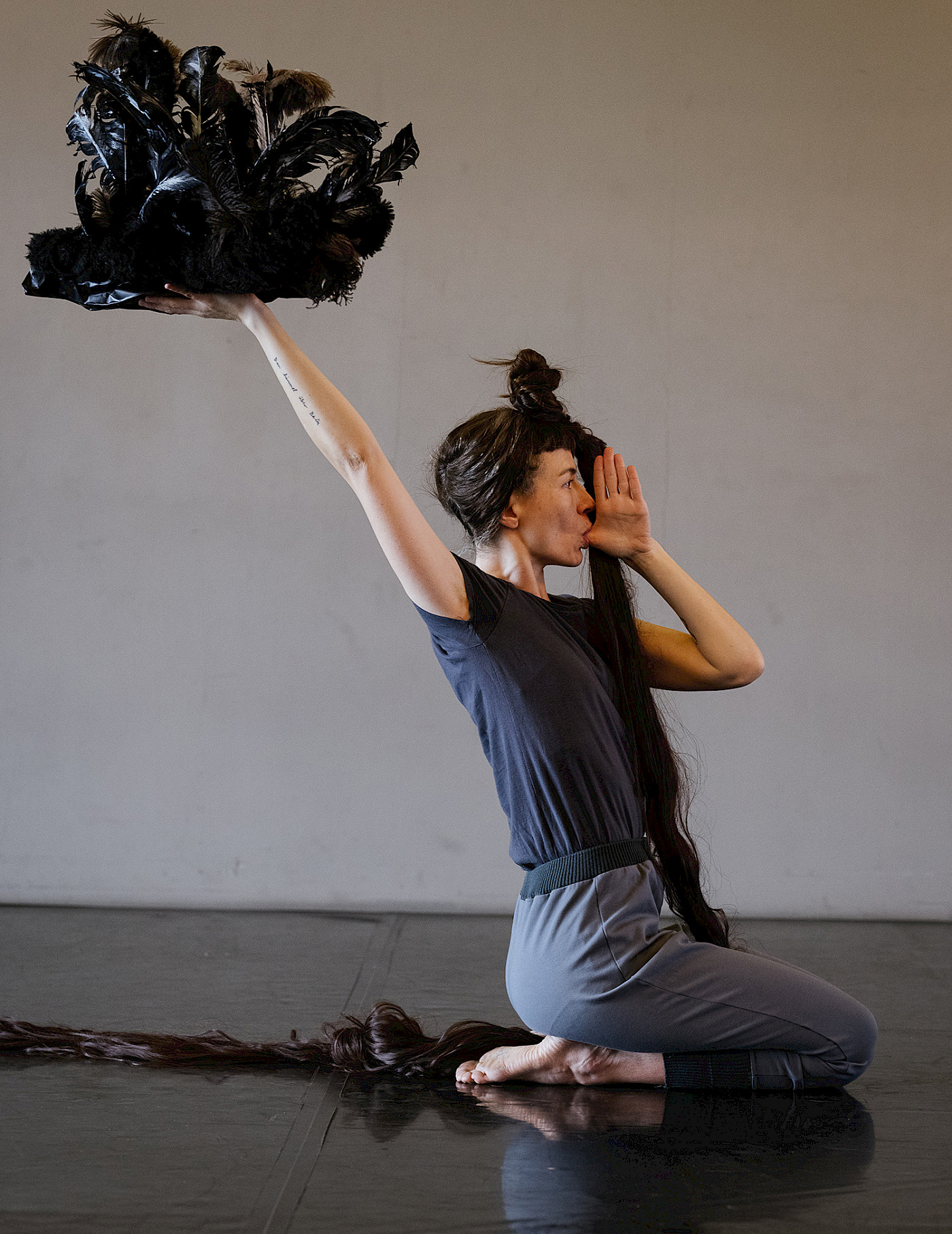 Dancer kneels on the floor, with her arm stretched out she holds a feather sculpture upwards, she has the thumb of her other hand in her mouth.