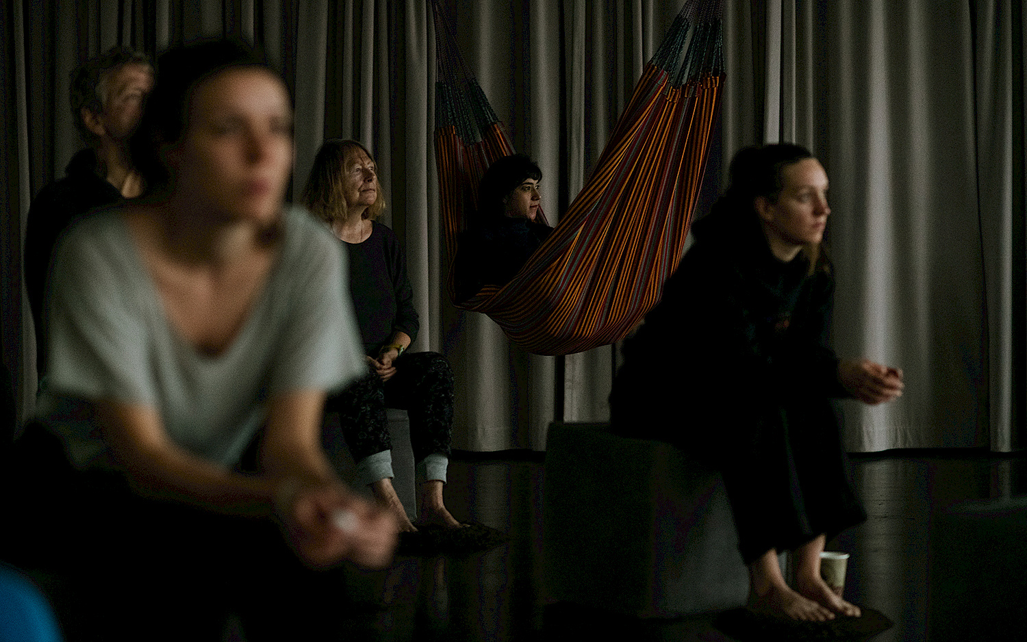 Several people are sitting in the twilight on stools or in hammocks. They are barefoot, with their feet on small piles of earth.