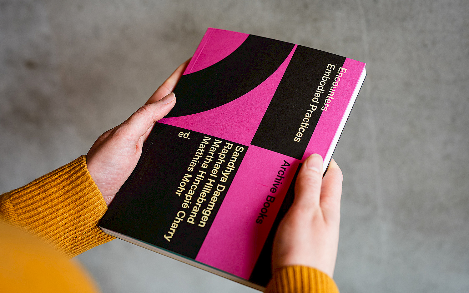 A person is holding the book "Encounters - Embodied Practices", the cover is graphically designed and black and pink.