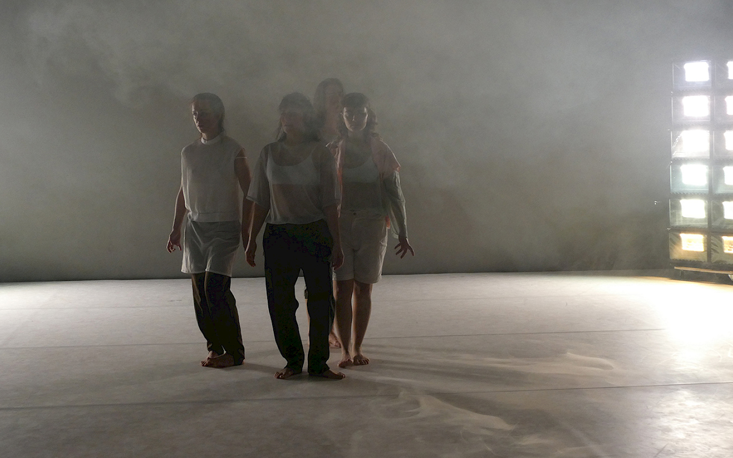 Vier Tänzerinnen stehen nah beieinander, die Bühne ist vernebelt, im Hintergrund leuchtet eine Lichinstallation hell.