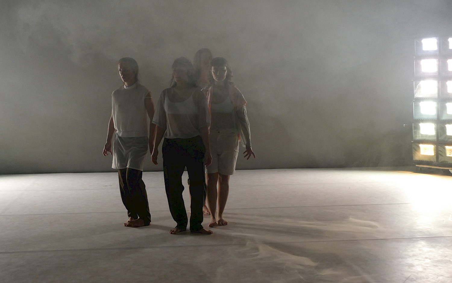 Four dancers stand close together, the stage is fogged, a light installation shines brightly in the background.