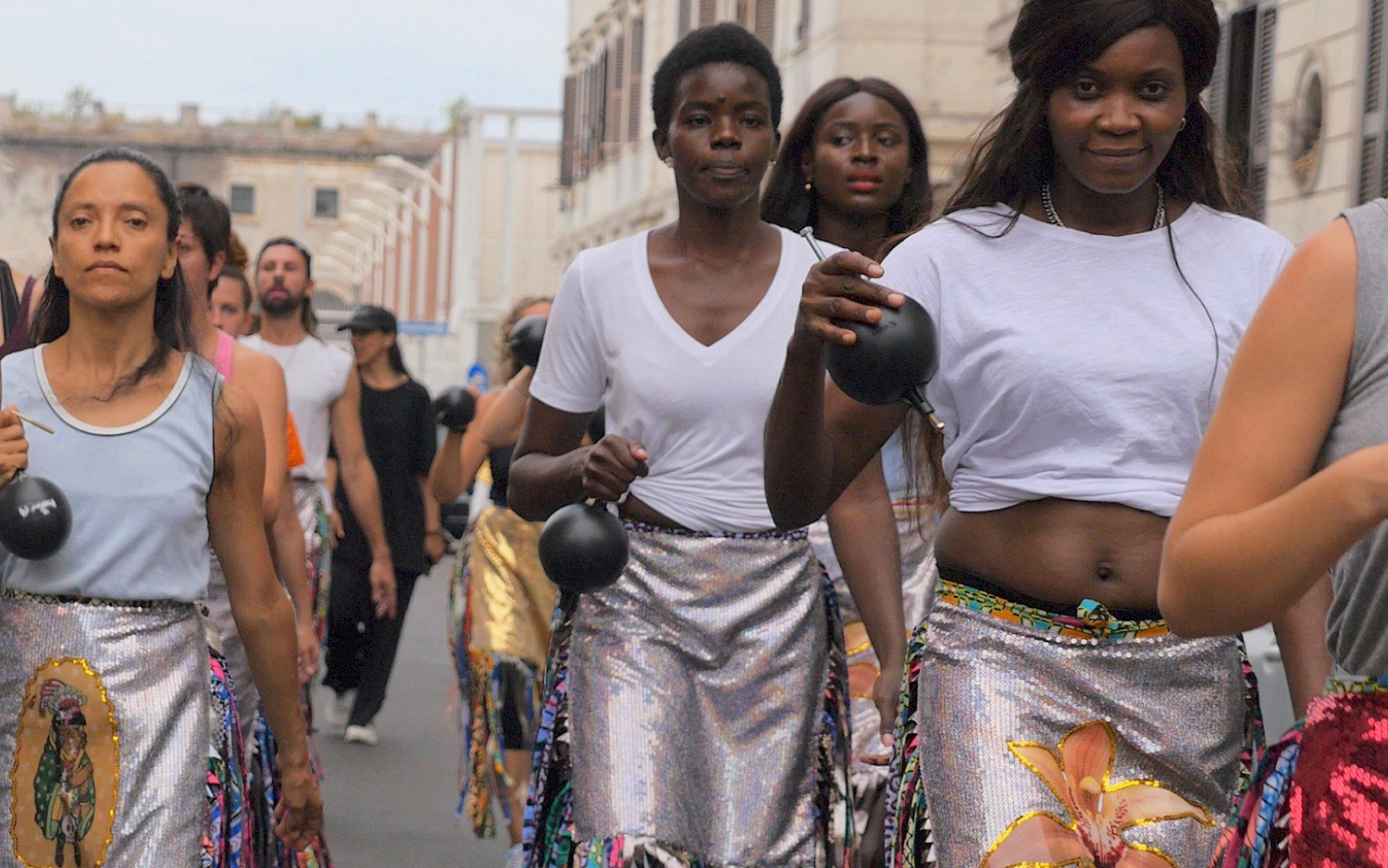 A group of dancers in costume walk down a street, holding heavy balls in their hands.