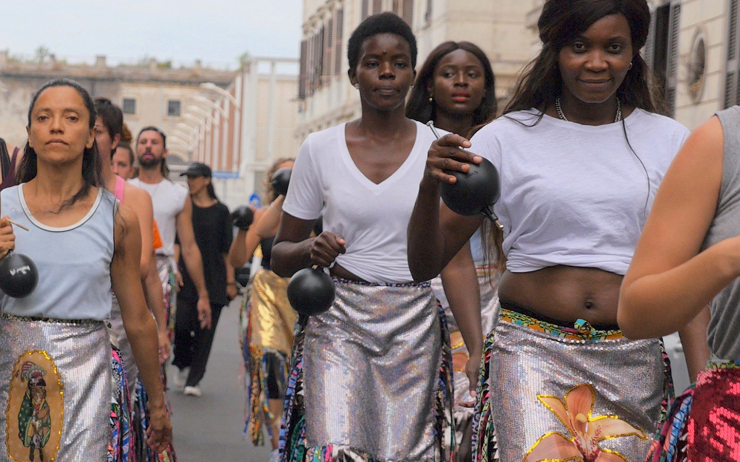 A group of dancers strides down a street, holding heavy bullets in their hands.
