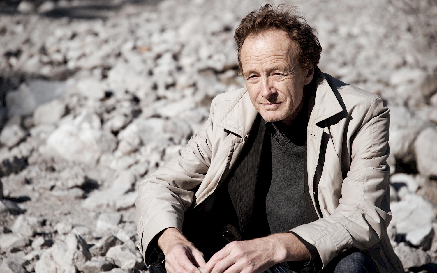 Der Komponist Beat Furrer sitzend in heller Jacke inmitten einer steinigen Landschaft.