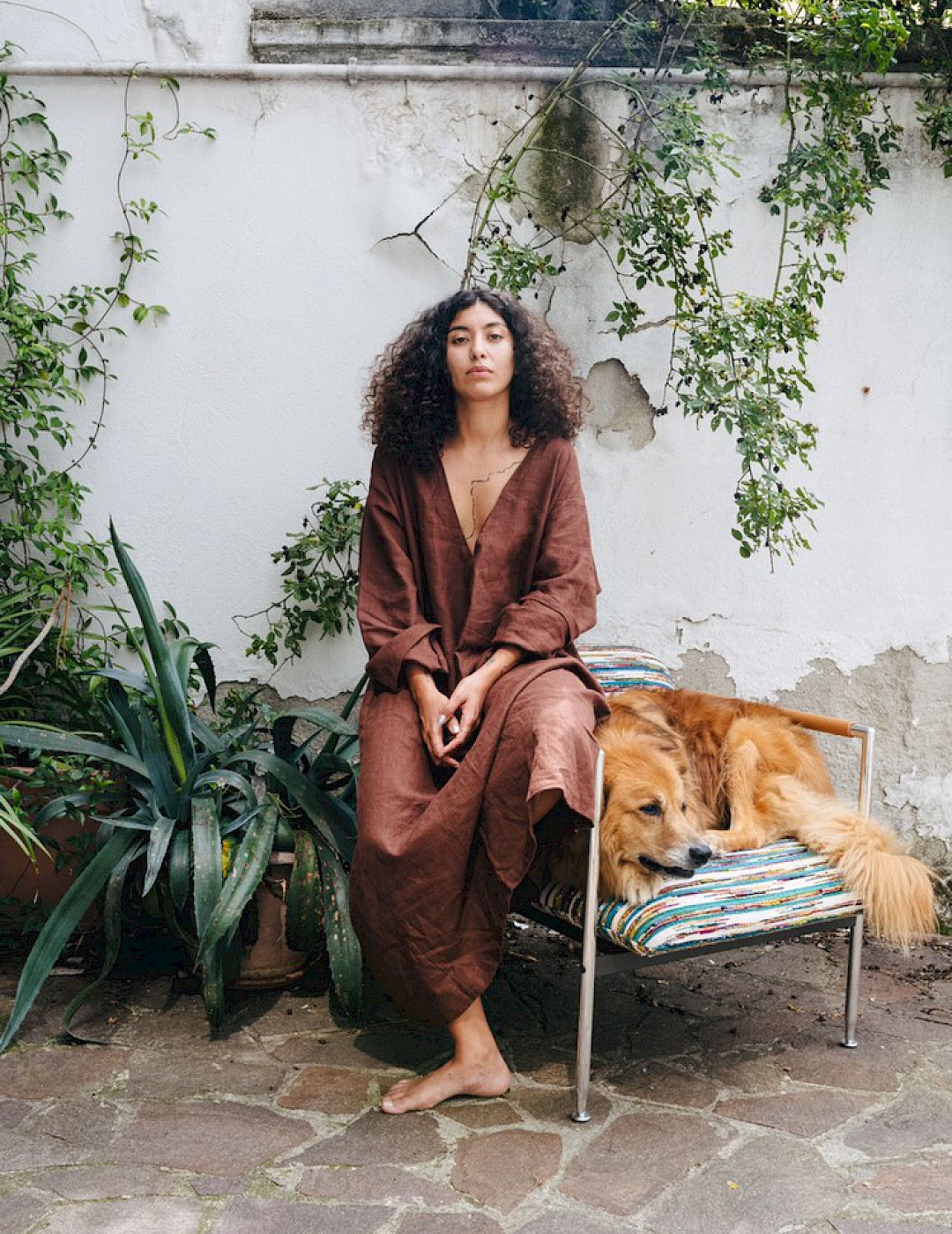 Frontales Ganzkörperportrait von Leila Bencharnia vor einer Mauer mit Pflanzen. Sie sitzt neben einem Hund auf der Armlehne eines Sessels.
