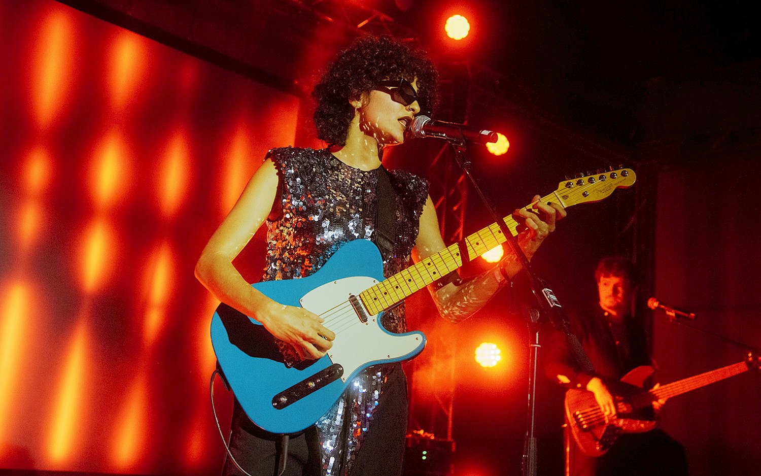 Rasha Nahas mit Sonnenbrille und E-Gitarre auf einer rot beleuchteten Bühne